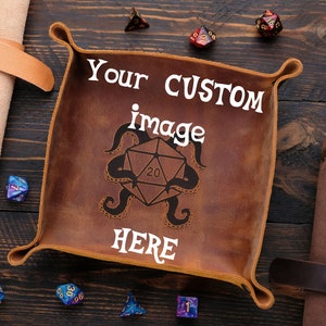 May include: A brown leather dice tray with a stitched border and a white inscription that reads "Your CUSTOM image HERE". The tray is surrounded by various colored dice on a wooden surface.