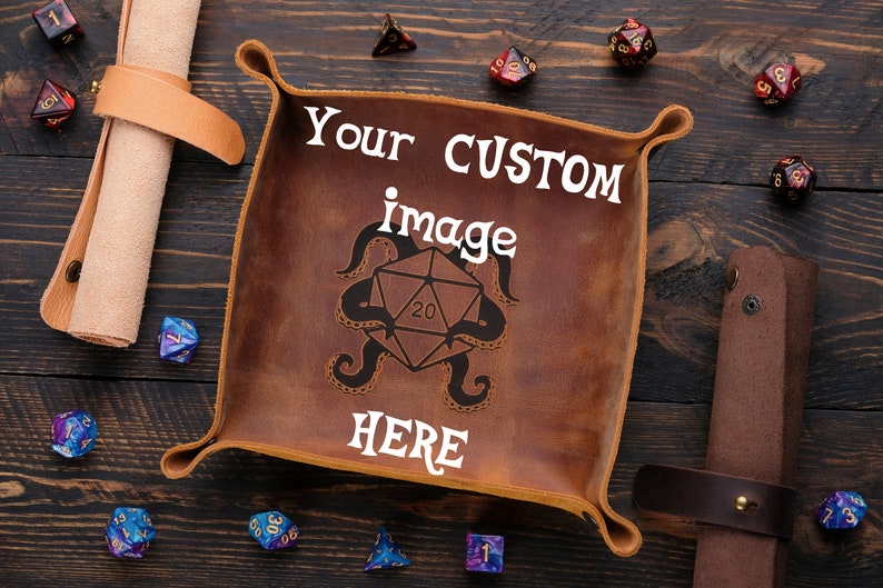 May include: A brown leather dice tray with a stitched border and a white inscription that reads "Your CUSTOM image HERE". The tray is surrounded by various colored dice on a wooden surface.