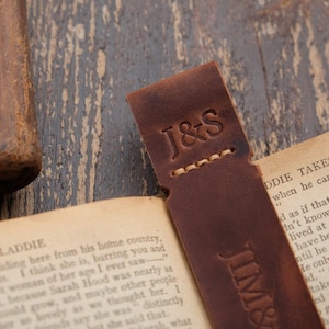 Pode incluir: Um marcador de p&aacute;gina de couro marrom com as iniciais "J&S" gravadas na frente. O marcador est&aacute; sobre um livro aberto com o texto "LADDIE TAKES THE PLUNGE" vis&iacute;vel.