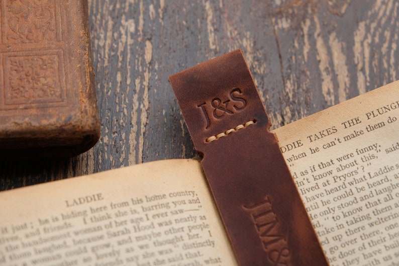 Pode incluir: Um marcador de p&aacute;gina de couro marrom com as iniciais "J&S" gravadas na frente. O marcador est&aacute; sobre um livro aberto com o texto "LADDIE TAKES THE PLUNGE" vis&iacute;vel.