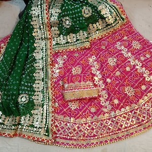 May include: A green and pink traditional Indian outfit with gold embroidery and embellishments. The outfit includes a skirt, a dupatta, and a matching blouse.