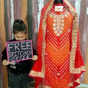 Puede incluir: Un salwar kameez rojo y naranja con estampado blanco y dorado. El atuendo está en un maniquí y tiene un ribete rosa y dorado. Un niño sostiene un cartel que dice "OFERTA DE COSTURA GRATUITA".