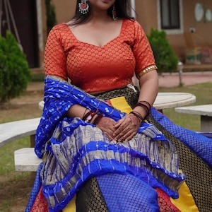 May include: An orange, patterned top with short sleeves and a low neckline. The outfit includes a blue and gold patterned shawl and a multi-colored skirt with red, yellow, black, and blue sections. The ensemble is a traditional Indian style.