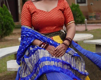 May include: An orange, patterned top with short sleeves and a low neckline. The outfit includes a blue and gold patterned shawl and a multi-colored skirt with red, yellow, black, and blue sections. The ensemble is a traditional Indian style.