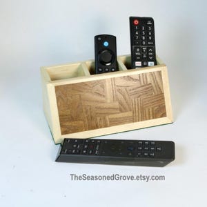 May include: A wooden remote control organiser with three slots. The organiser has a light wood frame and a dark wood inlay. Three remotes are in the slots, including a black remote with buttons, a black remote with a circular button, and a black remote with a directional pad.