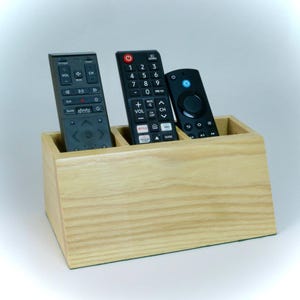 May include: A light-colored wooden remote control holder with three compartments, each holding a black remote. The holder is made of light-colored wood and has a simple, rectangular design. The remotes have various buttons and labels.