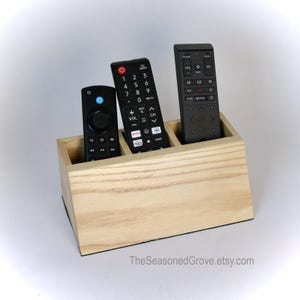 May include: A wooden remote control organizer with three slots. The organizer is made of light wood and has a natural finish. Three black remote controls are in the slots.