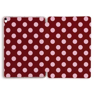 May include: A tablet case with a burgundy background and a pattern of evenly spaced, light pink polka dots. The case is open, revealing the interior and exterior design. The polka dots are consistent in size and spacing.