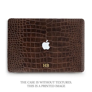 Può includere: Una custodia per laptop marrone scuro con un motivo stampato a pelle di coccodrillo, un logo Apple bianco ritagliato al centro e le iniziali HB color oro stampate vicino al bordo inferiore. La superficie è liscia e priva di texture.