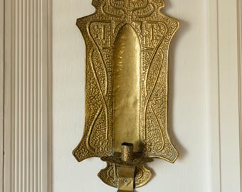 May include: An antique brass wall sconce with an Art Nouveau design. The sconce features a textured surface and intricate embossed patterns. It has a candle holder at the bottom and is mounted on a white wall. The sconce is approximately 12 inches tall.