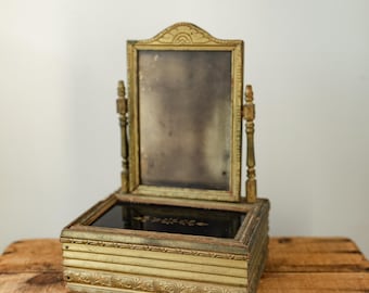 May include: An antique vanity mirror with a rectangular frame and a small storage box. The mirror and box are painted gold with decorative trim. The mirror is supported by two ornate posts. The box has a dark interior with a floral design.