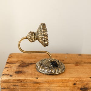 May include: An antique brass wall sconce with a decorative, fluted shade and a curved arm. The sconce is mounted on a round base with an ornate design. The fixture is made of brass and is set against a wooden surface.