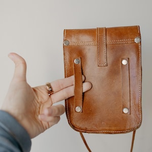 May include: A brown leather crossbody bag with a flap closure and silver rivets. The bag has a long strap and a small, rectangular shape. The bag is held by a hand.