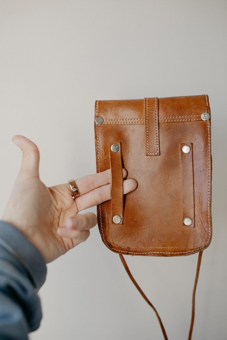 May include: A brown leather crossbody bag with a flap closure and silver rivets. The bag has a long strap and a small, rectangular shape. The bag is held by a hand.