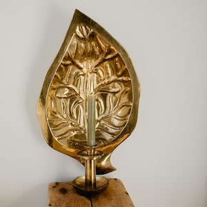 Peut inclure: Un bougeoir mural en forme de feuille en laiton avec un porte-bougie. Le bougeoir présente un motif texturé et détaillé et contient une bougie verte. Le porte-bougie est rond et repose sur une surface en bois.