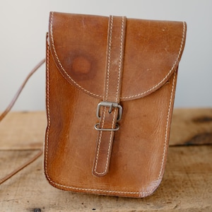 May include: A brown leather crossbody bag with a flap closure and a buckle. The bag has white stitching and a long strap. The leather has a worn appearance. The bag is sitting on a wooden surface.