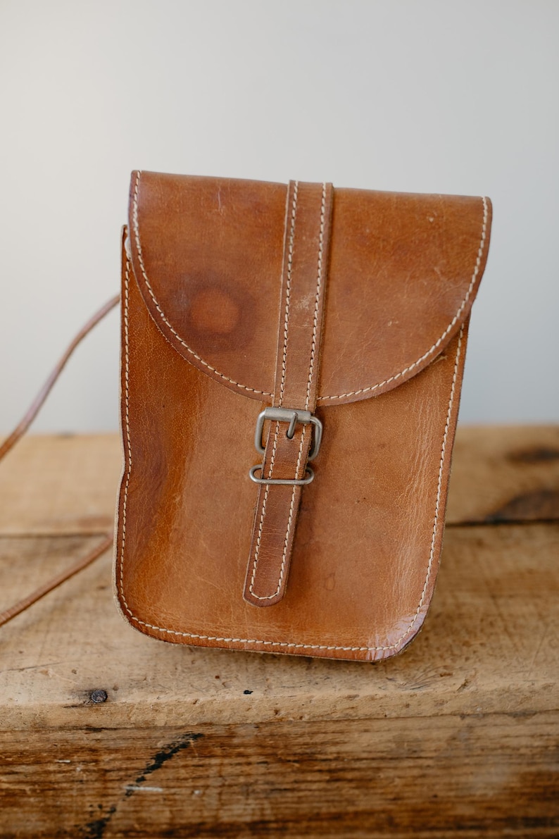 May include: A brown leather crossbody bag with a flap closure and a buckle. The bag has white stitching and a long strap. The leather has a worn appearance. The bag is sitting on a wooden surface.