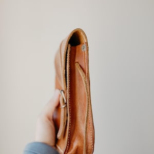 May include: A side view of a small, brown leather crossbody bag. The bag has a flap closure with a silver buckle and a long, thin strap. The leather appears to be worn and has visible stitching along the edges.