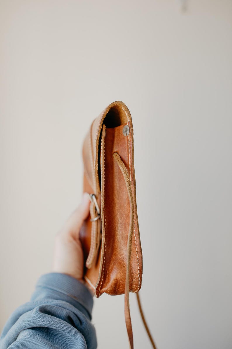 May include: A side view of a small, brown leather crossbody bag. The bag has a flap closure with a silver buckle and a long, thin strap. The leather appears to be worn and has visible stitching along the edges.