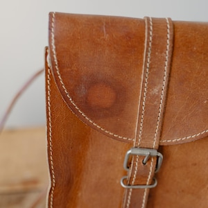 May include: Close-up of a brown leather satchel bag. The bag features a flap closure with a buckle and a strap. White stitching accents the edges of the bag and strap. The leather has a worn appearance.