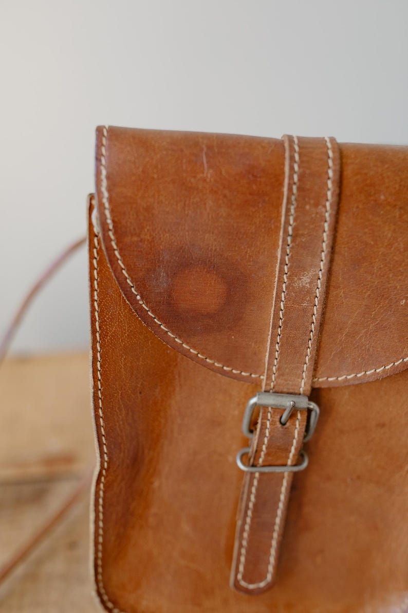 May include: Close-up of a brown leather satchel bag. The bag features a flap closure with a buckle and a strap. White stitching accents the edges of the bag and strap. The leather has a worn appearance.