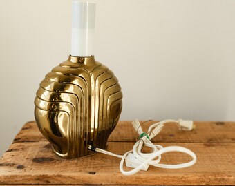 May include: A gold-colored table lamp with a spherical base and a white, rectangular shade. The lamp has a unique, layered design and a white power cord. The lamp is sitting on a wooden surface.