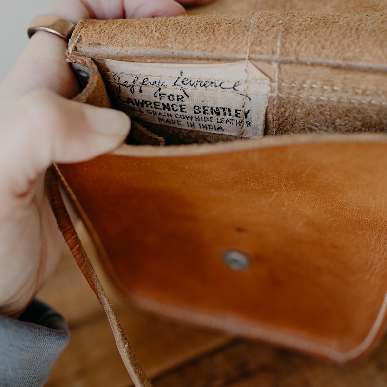 May include: Close-up of a tan leather bag interior. A tag reads "Jeffery Lawrence for Lawrence Bentley, Full Grain Cow Hide Leather, Made in India." The bag has a snap closure and a strap.