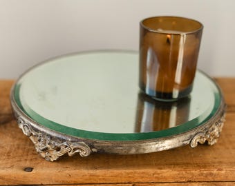 May include: A round, mirrored tray with an ornate silver-colored metal edge. A brown glass candle holder sits on the tray. The tray is on a wooden surface.