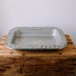 Pode incluir: Uma bandeja retangular de metal prateado com uma borda floral decorativa. A bandeja está vazia e repousa sobre uma caixa de madeira rústica. O design da bandeja apresenta um padrão repetido de folhas e vinhas ao redor da borda.