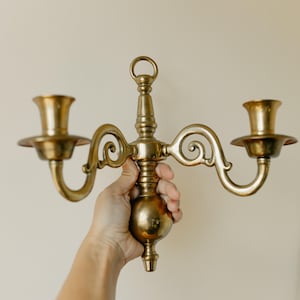 May include: A polished brass wall sconce featuring two candle holders with curved arms extending from a central vertical post. The fixture includes a circular ring at the top for mounting and a rounded, weighted base at the bottom, all in gold.