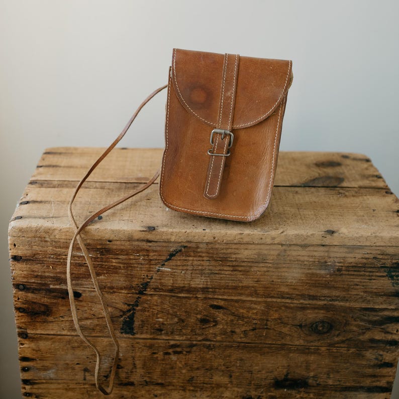 May include: A brown leather crossbody bag with a flap closure and a buckle. The bag has a long, thin strap and is sitting on a wooden crate. The leather has visible wear and tear, giving it a vintage look.