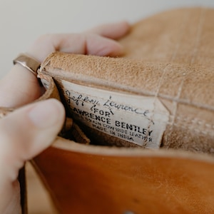 May include: Close-up of a tan leather bag interior. A label reads "Jeffrey Lawrence for Lawrence Bentley, Grain Cow Hide Leather, Made in India." The bag is open, revealing the textured leather and stitching.