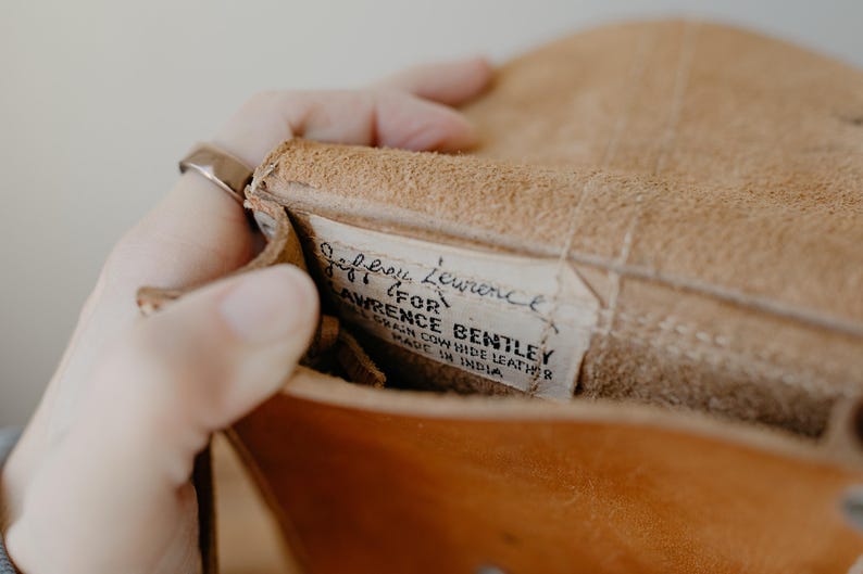 May include: Close-up of a tan leather bag interior. A label reads "Jeffrey Lawrence for Lawrence Bentley, Grain Cow Hide Leather, Made in India." The bag is open, revealing the textured leather and stitching.