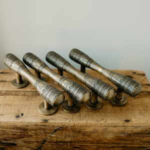 May include: Set of four antique bronze door pulls with intricate geometric patterns. Each pull features a cylindrical handle and a circular base, resting on a weathered wooden surface. The pulls are approximately 6 inches long.