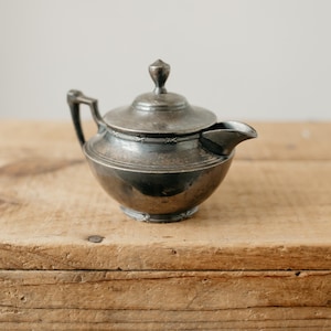 Peut inclure: Un pot à crème en métal argenté terni avec couvercle, doté d'une anse courbée, d'un petit bec verseur et d'un corps arrondi. La surface présente des signes d'usure et d'oxydation, avec une bande décorative entourant le couvercle et le bord.