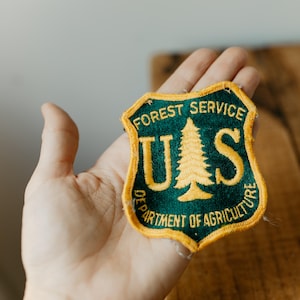 May include: A shield-shaped embroidered patch featuring a dark green background with a yellow border. The text FOREST SERVICE is arched at the top, with DEPARTMENT OF AGRICULTURE at the bottom. A yellow pine tree sits between the letters U and S.