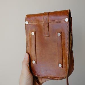 May include: A brown leather crossbody bag with a flap closure and a long strap. The bag features silver-colored rivets and visible stitching. The bag is held by a hand.