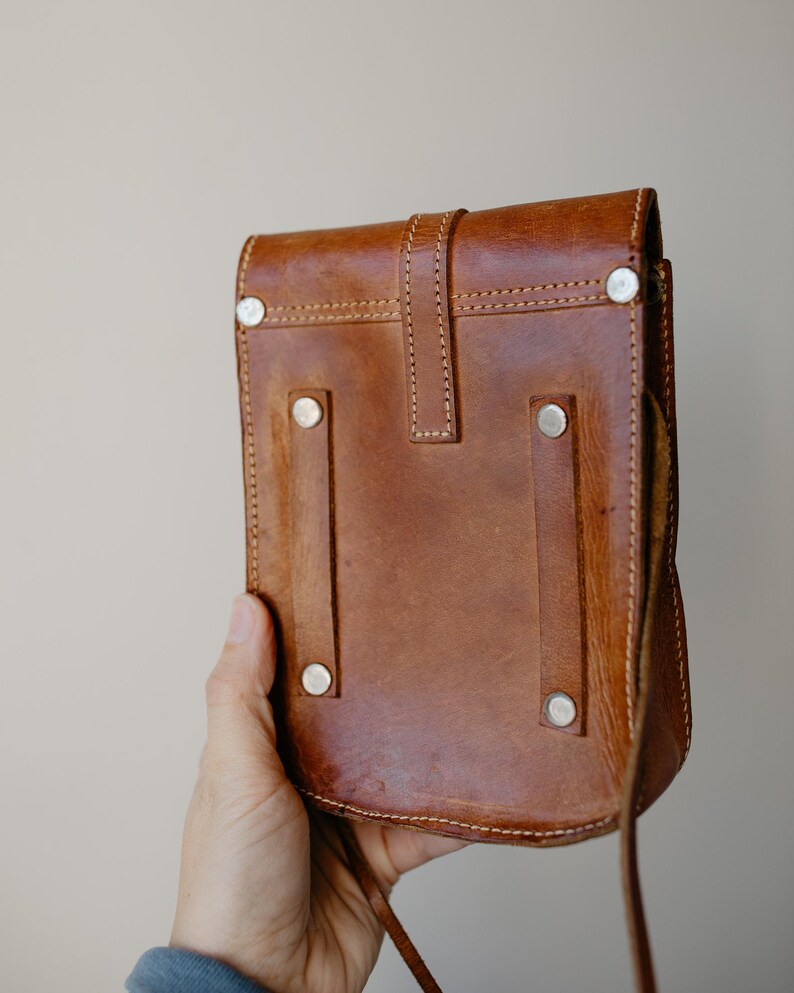 May include: A brown leather crossbody bag with a flap closure and a long strap. The bag features silver-colored rivets and visible stitching. The bag is held by a hand.