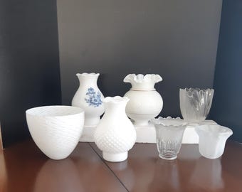 May include: A collection of seven white and clear glass lampshades. The lampshades are in various shapes and sizes, including a ruffled shade, a textured shade, and a clear glass shade.