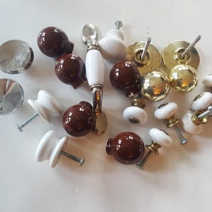 May include: Assortment of brown and white ceramic cabinet knobs and pulls, including a single white ceramic pull with a metal handle. The knobs and pulls have a variety of styles and finishes.