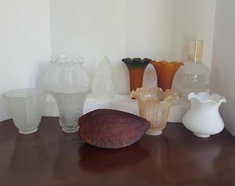 May include: A collection of eight glass lampshades in various shapes and sizes. The lampshades are made of frosted glass and have a variety of designs, including a floral pattern, a scalloped edge, and a simple, rounded shape. The lampshades are arranged on a wooden surface.