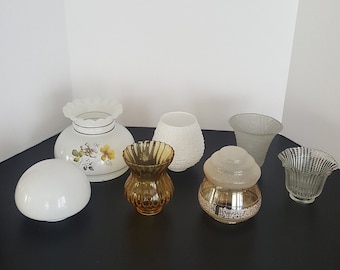 May include: A collection of seven white and clear glass lampshades, including a floral patterned shade, a textured shade, and a clear shade with a scalloped edge. The lampshades are arranged on a black surface.