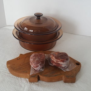Puede incluir: Un juego de dos platos para hornear de vidrio marrón con tapas apiladas una encima de la otra. Los platos están sobre una superficie blanca. Dos filetes crudos están sobre una tabla de cortar de madera con forma de cerdo.