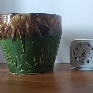 Peut inclure: Un pot en céramique émaillée vert et brun avec un motif géométrique. Le pot est posé sur une surface en bois à côté d'un réveil blanc.