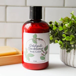 May include: A red body lotion bottle with a black cap and a white label featuring the text "Patchouli Sandalwood Body Lotion" and botanical illustrations. The bottle contains 8 oz of lotion. A bar of soap and a small plant are also in the image.