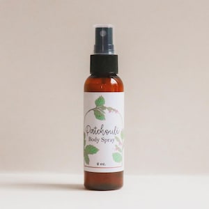 May include: An amber-colored glass bottle of body spray with a black plastic spray nozzle. The white label features a green leaf illustration and the text Patchouli Body Spray 2 oz. The bottle is cylindrical with a smooth, clear plastic cap.