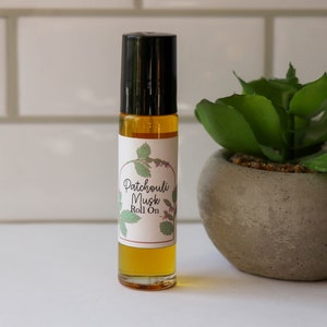 May include: A clear glass roll-on bottle with a black cap and a white label that reads "Patchouli Musk Roll On". The label has a green and brown illustration of a plant.