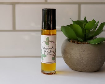 May include: A clear glass roll-on bottle with a black cap and a white label that reads "Patchouli Musk Roll On". The label has a green and brown illustration of a plant.