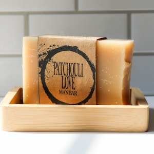 May include: A bar of soap in a bamboo soap dish. The soap is wrapped in brown paper with the text "Patchouli Love Manbar" printed on it.