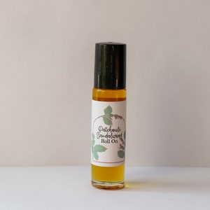 May include: A small glass bottle with a black roll-on top. The bottle is filled with a yellow liquid and has a white label with the text "Patchouli Sandalwood Roll On".